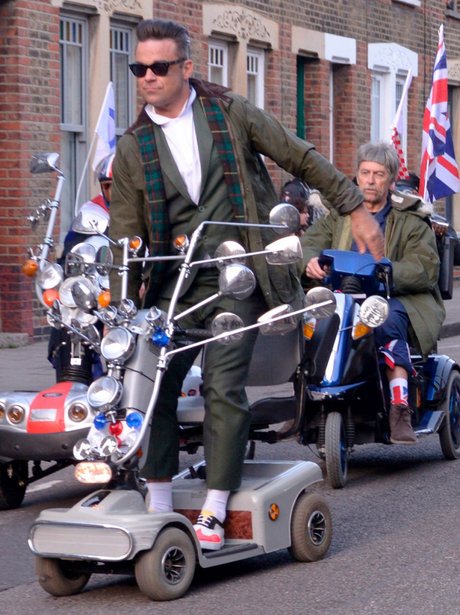 Robbie Williams stanidn up on Mobility Scooter wearing a green jacket and sunglasses